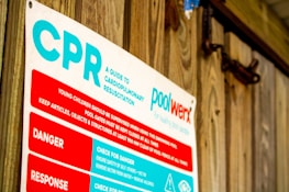 A sign detailing cardiopulmonary resuscitation (CPR) instructions, positioned against a wooden wall. The text includes warnings and response tips and highlights the importance of supervision around pools.