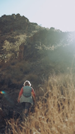 A person is hiking up a rocky, arid hill surrounded by dry grass and sparse desert vegetation. The sun is bright, casting a strong light that washes over the scene, creating a serene and adventurous atmosphere.