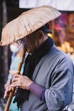 A person wearing traditional attire holds a bamboo flute under a woven straw hat. The scene suggests a street setting with blurred background elements and another individual visible but out of focus.