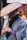 A person wearing traditional attire holds a bamboo flute under a woven straw hat. The scene suggests a street setting with blurred background elements and another individual visible but out of focus.