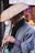 A person wearing traditional attire holds a bamboo flute under a woven straw hat. The scene suggests a street setting with blurred background elements and another individual visible but out of focus.