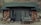 A rustic cabin with a stone exterior and a green shingle roof located in a natural setting. The facade includes a wooden door and windows with curtains. Hiking poles and shoes are placed by the door, indicating outdoor activity. A bench labeled 'Phantom Ranch' is seen beside the entrance, along with a fire extinguisher mounted on the wall.