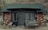 A rustic cabin with a stone exterior and a green shingle roof located in a natural setting. The facade includes a wooden door and windows with curtains. Hiking poles and shoes are placed by the door, indicating outdoor activity. A bench labeled 'Phantom Ranch' is seen beside the entrance, along with a fire extinguisher mounted on the wall.