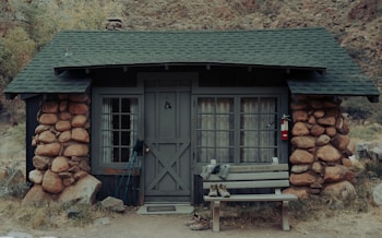 A rustic cabin with a stone exterior and a green shingle roof located in a natural setting. The facade includes a wooden door and windows with curtains. Hiking poles and shoes are placed by the door, indicating outdoor activity. A bench labeled 'Phantom Ranch' is seen beside the entrance, along with a fire extinguisher mounted on the wall.
