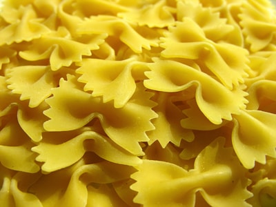 A close-up view of uncooked bow-tie pasta with a smooth, yellow surface and distinct shape resembling butterfly wings. The noodles are neatly piled together, showcasing their unique form with ridged edges.