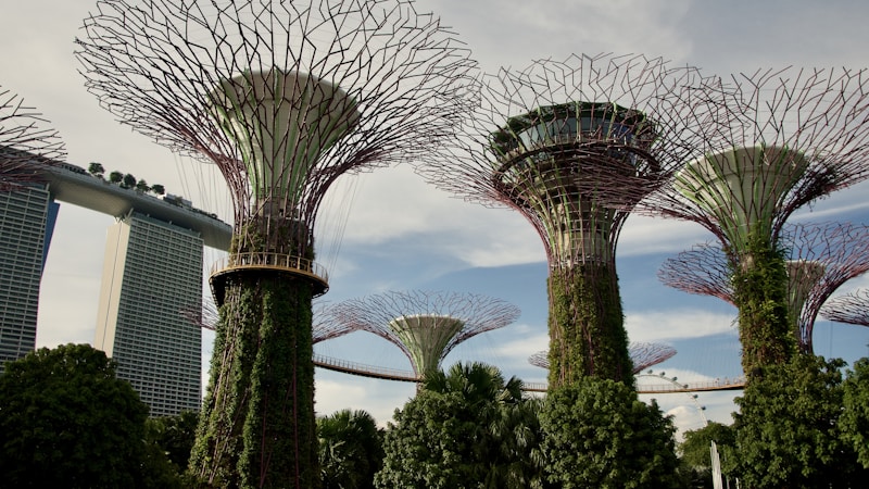 Vibrant garden structures resembling giant trees are covered in greenery and plants. A modern architectural building with a boat-like top is visible in the background, suggesting a futuristic and eco-friendly urban design.