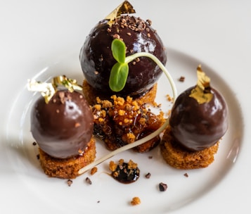 A gourmet dessert consisting of three dark chocolate spheres with gold leaf accents, placed on top of small round cake pieces. Between the chocolate pieces, there is a garnish of crumbled bits and a decorative plant sprout. The overall presentation is elegant and artistic.