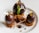 A gourmet dessert consisting of three dark chocolate spheres with gold leaf accents, placed on top of small round cake pieces. Between the chocolate pieces, there is a garnish of crumbled bits and a decorative plant sprout. The overall presentation is elegant and artistic.