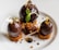 A gourmet dessert consisting of three dark chocolate spheres with gold leaf accents, placed on top of small round cake pieces. Between the chocolate pieces, there is a garnish of crumbled bits and a decorative plant sprout. The overall presentation is elegant and artistic.