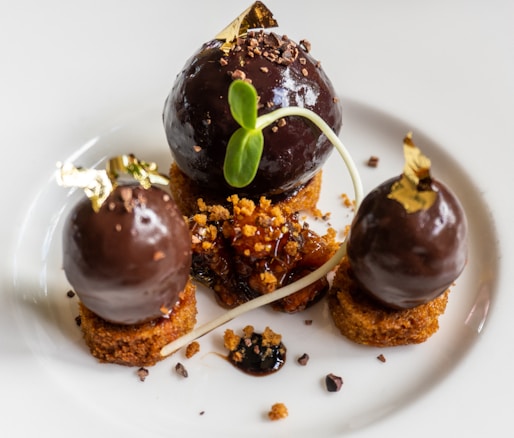 A gourmet dessert consisting of three dark chocolate spheres with gold leaf accents, placed on top of small round cake pieces. Between the chocolate pieces, there is a garnish of crumbled bits and a decorative plant sprout. The overall presentation is elegant and artistic.