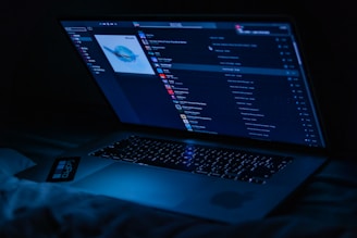 A laptop displaying a music player interface is illuminated in a dark setting, casting a blue glow. The screen shows a playlist with album art and various song titles.