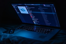 A laptop displaying a music player interface is illuminated in a dark setting, casting a blue glow. The screen shows a playlist with album art and various song titles.
