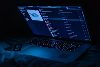 A laptop displaying a music player interface is illuminated in a dark setting, casting a blue glow. The screen shows a playlist with album art and various song titles.