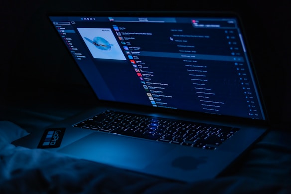 A laptop displaying a music player interface is illuminated in a dark setting, casting a blue glow. The screen shows a playlist with album art and various song titles.