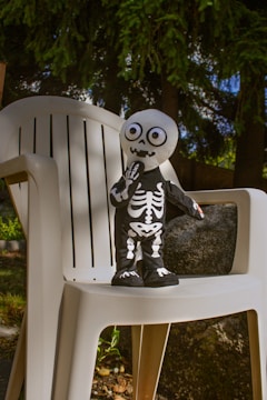 A small skeleton-themed doll stands on a plastic chair outdoors, surrounded by greenery. The doll is dressed in a black costume with white skeletal patterns and has oversized googly eyes. The background features trees and shrubs under natural daylight.