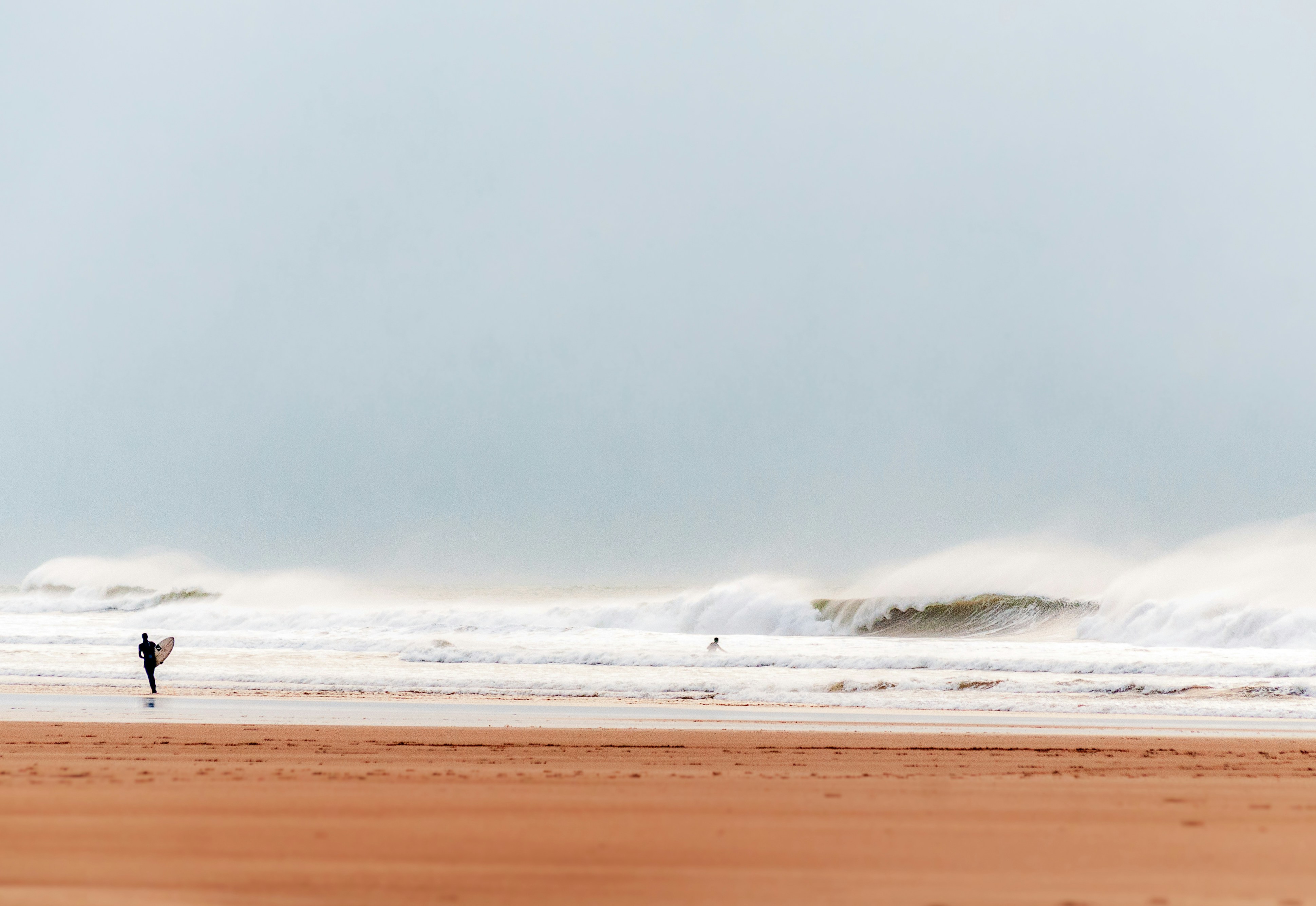 Strong offshore wind with large waves at Croyde Beach in Croyde Bay. Surfer with surf board in wetsuit walking along the sand.