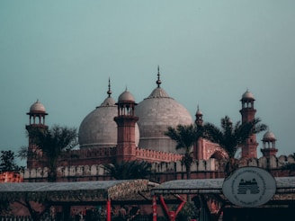 A grand historical mosque with large domes and minarets is surrounded by lush palm trees. The architectural design features intricate detailing on the walls and domes. The sky above is clear, adding a calming backdrop to the scene.