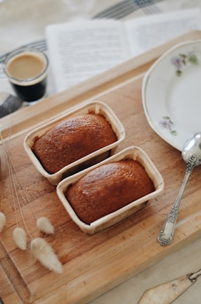 Two freshly baked loaves in small paper containers rest on a wooden cutting board. Beside them lies a silver spoon and a floral-decorated porcelain plate. To the side is a small glass cup filled with espresso, set next to an open book. Soft, fluffy beige grass stems artfully garnish the board.