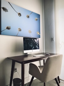 A modern workspace featuring a sleek, minimalist desk setup with a computer, keyboard, and mouse. Above the desk hangs a large canvas depicting palm trees stretching toward a clear blue sky. The room is well-lit with a soft, natural light from the side window, providing a calm and serene atmosphere.