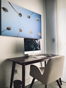 A modern workspace featuring a sleek, minimalist desk setup with a computer, keyboard, and mouse. Above the desk hangs a large canvas depicting palm trees stretching toward a clear blue sky. The room is well-lit with a soft, natural light from the side window, providing a calm and serene atmosphere.