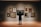 A person is standing in front of a wall displaying a variety of framed paintings in an art gallery. The artworks feature different subjects including landscapes and portraits, all arranged neatly on the wall. The person appears to be observing the central painting closely.