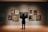 A person is standing in front of a wall displaying a variety of framed paintings in an art gallery. The artworks feature different subjects including landscapes and portraits, all arranged neatly on the wall. The person appears to be observing the central painting closely.