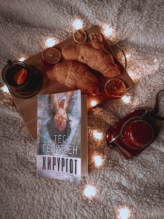 A cozy setup featuring a book written in Cyrillic script, placed on a soft textured surface surrounded by warm fairy lights. A wooden board holds two croissants and dried citrus slices, accompanied by a cup of tea with a slice of orange. A vintage-style camera is placed nearby.
