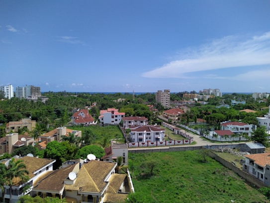 Aerial view of a residential area featuring a variety of buildings, including multi-story apartments and individual houses with red and brown roofs. The scene is lush with greenery, including trees and well-maintained lawns. The horizon shows a distant outline of the ocean under a bright blue sky with scattered clouds.