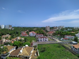 Aerial view of a residential area featuring a variety of buildings, including multi-story apartments and individual houses with red and brown roofs. The scene is lush with greenery, including trees and well-maintained lawns. The horizon shows a distant outline of the ocean under a bright blue sky with scattered clouds.