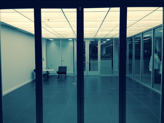 A modern, minimalistic office space with glass walls and doors. The room features a single black chair and a small table. The ceiling has a grid of white, illuminated panels, giving the room a bright, sterile feel. The floor is covered with large, dark tiles.