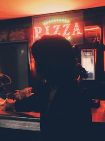 A dimly lit scene at a pizza shop, with a glowing neon sign that reads 'PIZZA' in the background. A silhouette of a person is visible at the counter, suggesting they might be making a transaction or waiting for an order. The atmosphere is warm with ambient lighting reflecting off surfaces.