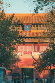 Ancient Chinese architecture with intricate designs in red and green adorns the entrance to a historical building. The building is partially obscured by lush green trees, and a few people and a bus are visible in the foreground.
