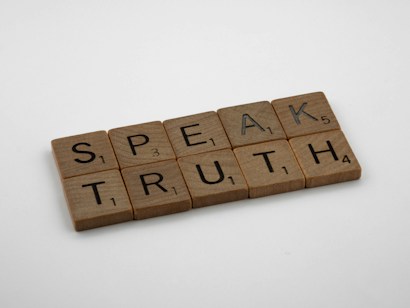 Wooden Scrabble tiles spell out the words 'SPEAK TRUTH' on a plain white background. Each tile displays a letter with a small number in the corner indicating its point value in the game.