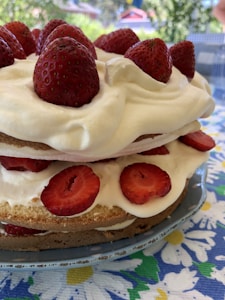 A delicious layer cake topped with whole strawberries and filled with whipped cream and sliced strawberries. The cake is set outdoors on a table with a colorful floral tablecloth, suggesting a bright and cheerful setting.