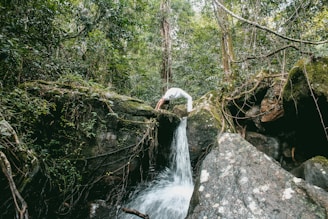 A person dressed in white is performing a backbend on a large rock in the middle of a lush forest. The setting includes a small waterfall with water cascading down into a pool below, surrounded by dense green foliage and moss-covered rocks.