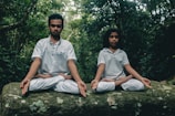 Two individuals sitting cross-legged on a large rock, both engaged in meditation. They wear white clothing and are surrounded by dense, green foliage, suggesting they are in a forest or jungle setting.