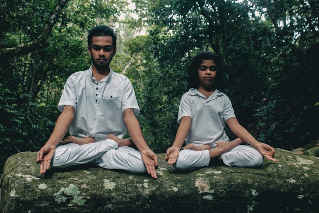Two individuals sitting cross-legged on a large rock, both engaged in meditation. They wear white clothing and are surrounded by dense, green foliage, suggesting they are in a forest or jungle setting.