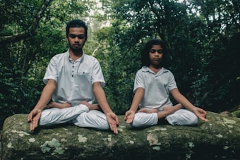 Two individuals sitting cross-legged on a large rock, both engaged in meditation. They wear white clothing and are surrounded by dense, green foliage, suggesting they are in a forest or jungle setting.