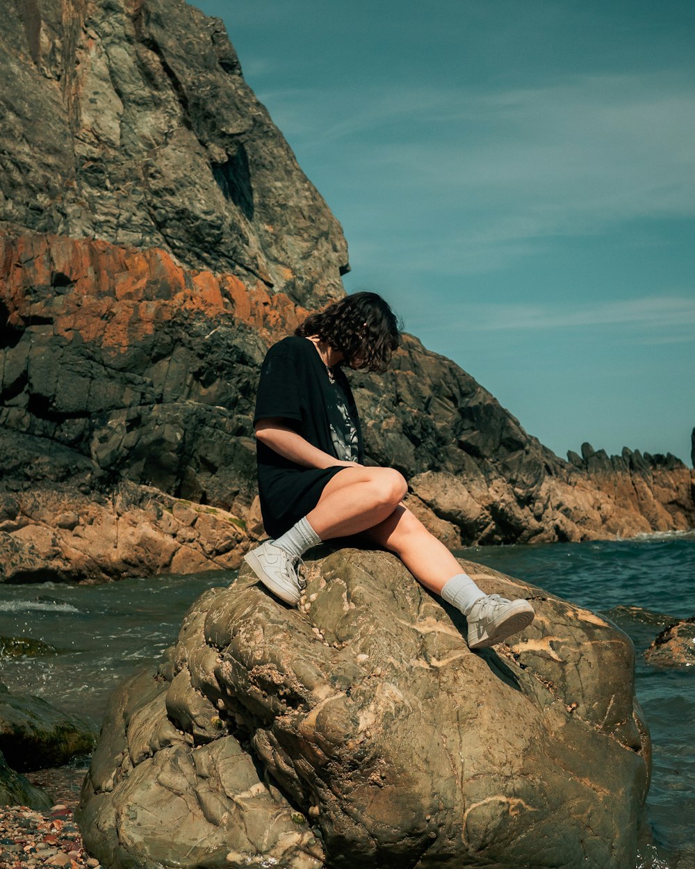 검은 셔츠와 흰색 반바지를 입은 여자가 낮에 수역 근처의 바위에 앉아 있습니다.