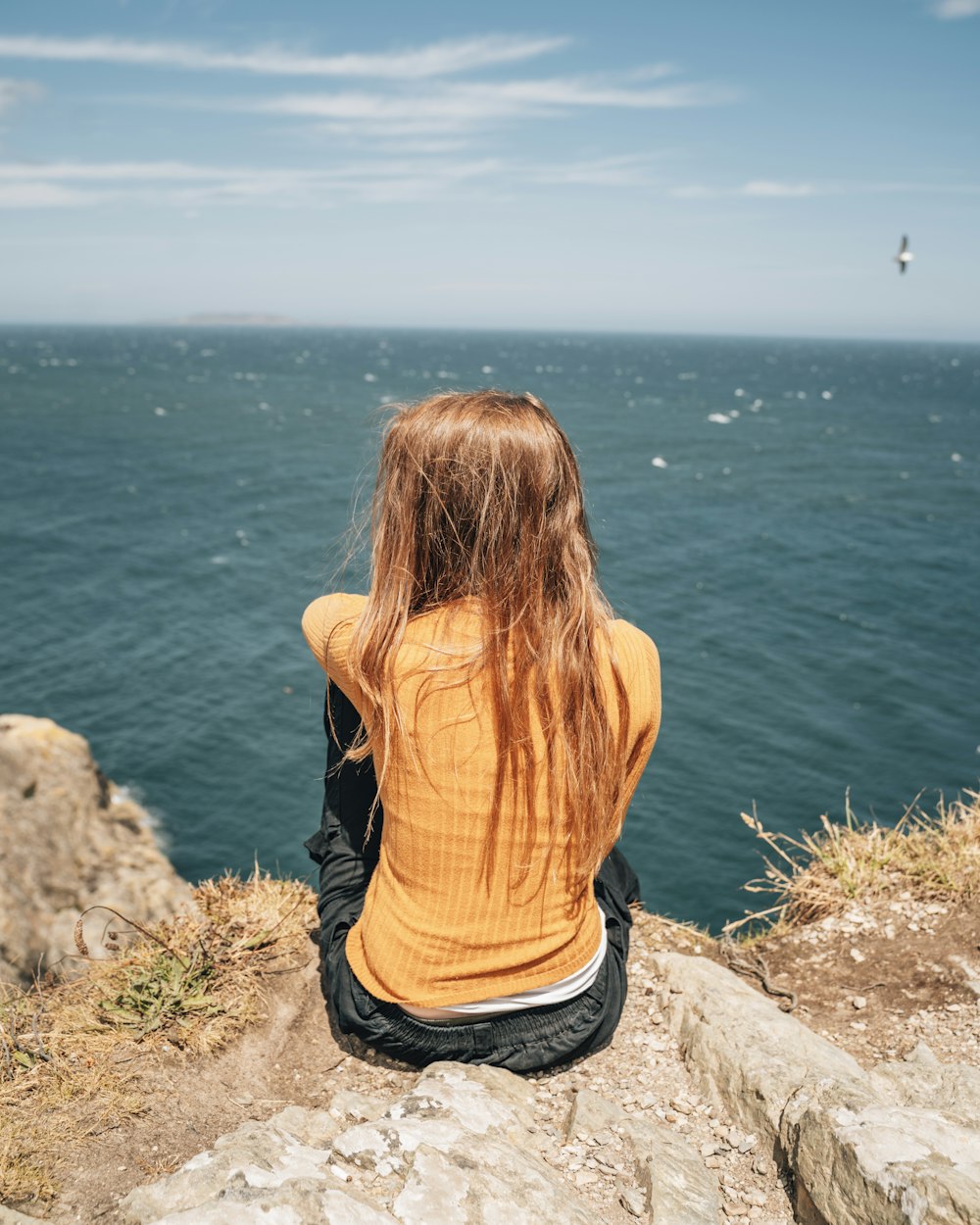 woman in yellow shirt and black pants sitting on rock near body of water during daytime