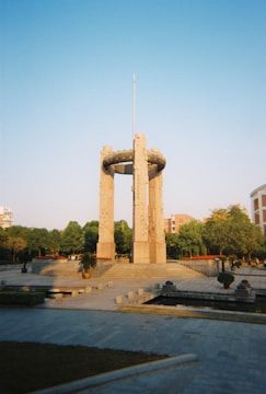 A tall stone monument features a central cylindrical structure supported by four large pillars. The monument is surrounded by well-maintained gardens and paved walkways. It is situated in an open area with trees and a few buildings in the background under a clear blue sky.