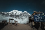 A stunning mountainous landscape with a snow-capped peak rising above a layer of clouds. In the foreground, there is a rustic open-air tea shop with a sign reading 'Mardi Tea Shop Viewpoint'. Wooden benches and tables are scattered around on the dirt ground, providing a spot to enjoy the breathtaking view.