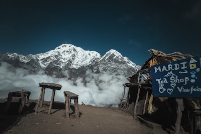 A stunning mountainous landscape with a snow-capped peak rising above a layer of clouds. In the foreground, there is a rustic open-air tea shop with a sign reading 'Mardi Tea Shop Viewpoint'. Wooden benches and tables are scattered around on the dirt ground, providing a spot to enjoy the breathtaking view.