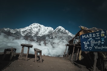 A stunning mountainous landscape with a snow-capped peak rising above a layer of clouds. In the foreground, there is a rustic open-air tea shop with a sign reading 'Mardi Tea Shop Viewpoint'. Wooden benches and tables are scattered around on the dirt ground, providing a spot to enjoy the breathtaking view.