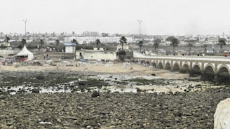 An expansive view of a rocky shoreline with a sandy beach area, bordered by a bridge with archways. People can be seen around the beach, some sitting under umbrellas. There are palm trees and a few small buildings in the background, with a line of parked cars along a road that runs parallel to the beach.