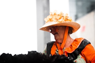 An elderly person is dressed in a colorful outfit with an orange blouse featuring a bright collar and black accents. They are wearing a straw hat adorned with orange flowers and are seen smiling in a candid moment.