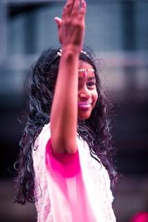 A person with long, dark, wavy hair adorned with a headband and bindi, dressed in a light pink attire, is captured with one arm raised. The background appears to be blurred, hinting at an outdoor setting.