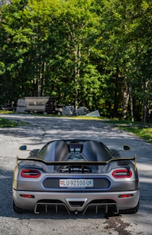 A sleek, high-performance sports car is parked on a quiet road surrounded by lush greenery. The rear view of the car highlights its aerodynamic design, prominent rear wing, and dual exhausts. The license plate indicates a location, and the car's surface reflects the sunlight, adding a dynamic sheen.