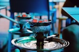 A hookah with glowing coals sits on a detailed metal tray. The setting appears casual, with blurred elements in the background and soft lighting that gives a blue tint.
