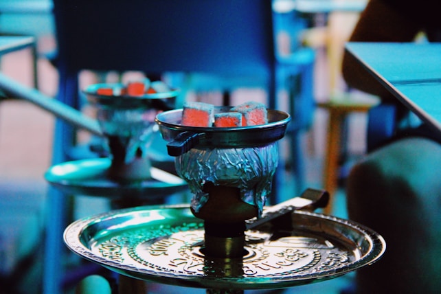 A hookah with glowing coals sits on a detailed metal tray. The setting appears casual, with blurred elements in the background and soft lighting that gives a blue tint.